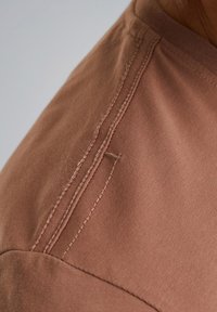 Hnědý bavlněný tričko se zaměřením na ramenní šev, s čistým šitím a hladkou texturou, s mírným leskem na povrchu látky.