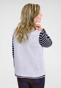 Grau strukturierte Weste über einem gestreiften Langarmoberteil in Schwarz und Weiß, kombiniert mit dunkelblauen Jeans. Die Weste hat einen abgerundeten Saum.