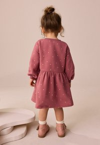 Peutermeisje staat, draagt een roze stippenjurk, witte sokken en roze laarzen, met haar in een rommelige bun, op een beige achtergrond.