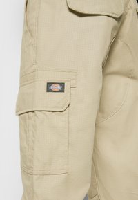 Pantalon cargo beige avec une poche latérale munie d'un rabat et d'une étiquette rectangulaire de la marque Dickies cousue sur la poche.