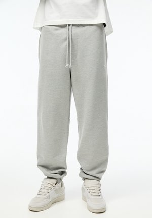 Pantalon de survêtement gris clair avec cordon, associé à des baskets blanches, porté par une personne debout devant un fond blanc uni.