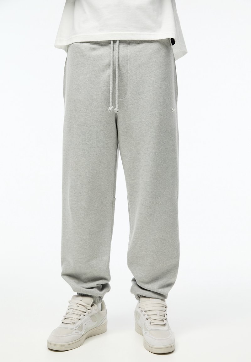 Pantalon de survêtement gris clair avec cordon, associé à des baskets blanches, porté par une personne debout devant un fond blanc uni.