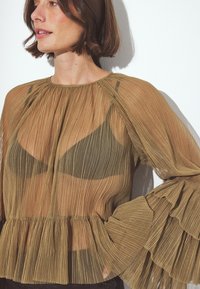 Blusa larga de manga larga, plisada y transparente en tonos terrestres, con un dobladillo con volantes, que presenta un escote alto y mangas de campana, superpuesta sobre un sujetador oscuro.