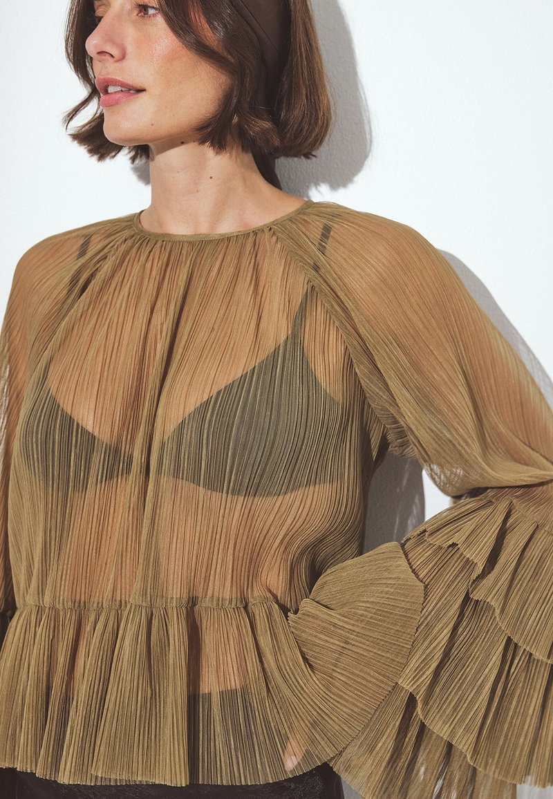 Blusa larga de manga larga, plisada y transparente en tonos terrestres, con un dobladillo con volantes, que presenta un escote alto y mangas de campana, superpuesta sobre un sujetador oscuro.