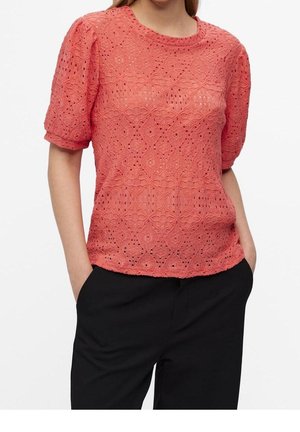Femme portant un chemisier en dentelle rose corail à manches courtes avec un col rond et un pantalon noir, mains dans les poches.