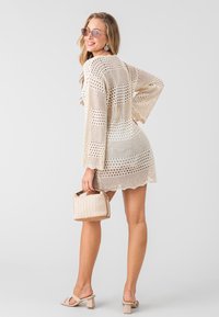 Beige haakjurk met een losse pasvorm en belmouwen, voorzien van open gebreide patronen; gecombineerd met een geweven handtas en hak sandalen.