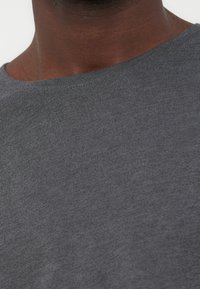 Κοντινό πλάνο του λαιμού και του πάνω μέρους του στήθους ενός ατόμου που φοράει απλό γκρι σκουρόχρωμο μπλουζάκι με στρογγυλή λαιμόκοψη, πάνω σε φωτεινό φόντο.