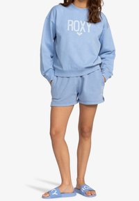 Felpa azzurra chiara con logo "ROXY", abbinata a pantaloni corti blu coordinati e sandali scivolati. Tessuto morbido, vestibilità rilassata, polsini a coste.