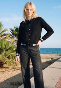 Young woman standing near beach palm trees, wearing a black varsity jacket with white stripes and black jeans under a clear blue sky.