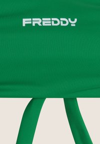 Vihreä urheilupaita, joka on valmistettu joustavasta kankaasta. Ominaisuuksiin kuuluu valkoinen "FREDDY" -logo ja koristeelliset solmut alareunassa. Sujuva rakenne, istuva muotoilu.