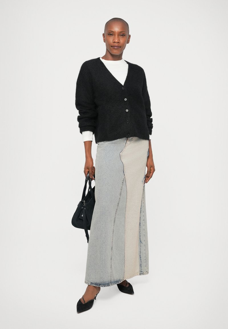 Cardigan nero in lana con scollatura a V e bottoni; abbinato a una gonna in denim a blocchi di colore lunga con cuciture visibili. Borsa nera, scarpe a punta.