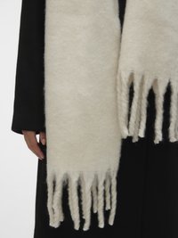 Henkilö, joka käyttää mustaa takkia ja päällä on suuri, pehmeä kermaan sävyinen huivi, jossa on paksut, kierretyt hapsut roikkumassa alaspäin.