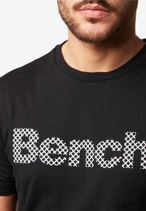 T-shirt en coton noir avec un logo "Bench" en motif en résille blanche. Le design à col rond et la texture douce sont des détails importants.