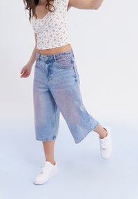 Joven con un top corto blanco de flores, pantalones culottes de mezclilla azul flojos y zapatillas blancas, de pie sobre una pierna contra un fondo liso.