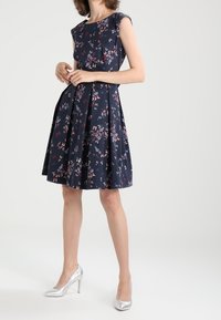 Marinblå klänning med blommigt mönster, kappärmar, figursydd överdel och utställd kjol. Silverfärgade högklackade skor fulländar outfiten.