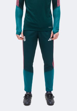 Uomo che indossa pantaloni da corsa sportivi blu petrolio scuro e rosa con una maglia abbinata, in piedi con le mani rilassate lungo i fianchi e tacchetti neri ai piedi.