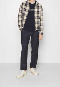 Chaqueta beige y azul marino a cuadros con cremallera sobre sudadera azul marino, pantalones oscuros y zapatos crema con cordones. Tejido suave, diseño informal.