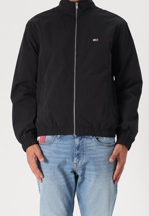 Homme portant une veste noire zippée avec des poignets élastiques et un jean bleu clair, les mains détendues le long du corps.