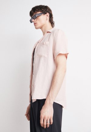Camicia rosa chiaro a maniche corte con bottoni in un tessuto texturizzato, dotata di taschino sul lato sinistro e una vestibilità rilassata. Indossata sopra una maglietta nera.
