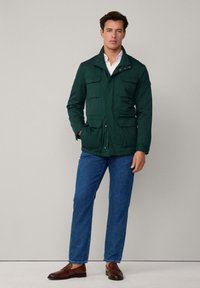 Grön, vattentät jacka med ståkrage, flera framfickor och dragkedja. I kombination med blå jeans och bruna slip-on skor.
