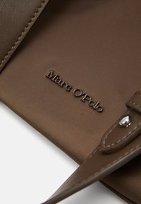Tissu texturé marron avec une marque Marc O’Polo. Comprend une sangle avec un bouton-pression en ton argent pour une fermeture sécurisée. Design minimaliste.