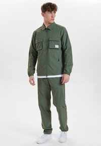 Zielona, lekka kurtka z dwoma kieszeniami na piersi, z zamkiem błyskawicznym i elastycznymi mankietami, zestawiona z pasującymi spodniami i białymi sneakerami.