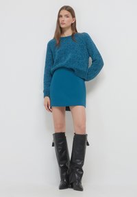 Strickpullover in Teal mit lockerer Passform, kombiniert mit einem taillierten mini Rock in Teal und kniehohen schwarzen Lederstiefeln mit Reißverschlüssen und Quasten.