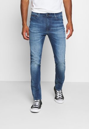 Mann trägt Slim-Fit blaue Jeans mit ausgewaschenen Details und schwarze High-Top-Sneaker mit weißen Schnürsenkeln, steht vor weißem Hintergrund.