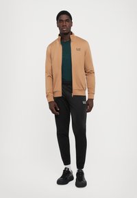 Jauns vīrietis stāv brūnā zip-up jakā, tumši zaļā krekla, melnās sporta biksēs, baltās zeķēs un melnās čībās pret vienkāršu baltu fonu.