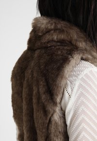 Braune Kunstpelzweste mit weicher Textur, ausgestattet mit einem hohen Kragen und einem mehrschichtigen Design. Detail eines weißen Hemdes mit Muster darunter.