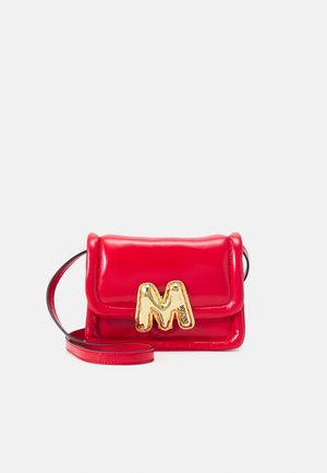 Mała błyszcząca czerwona skórzana torebka na ramię z złotą sprzączką w kształcie litery "M" i pasującym czerwonym paskiem na jasnym tle.