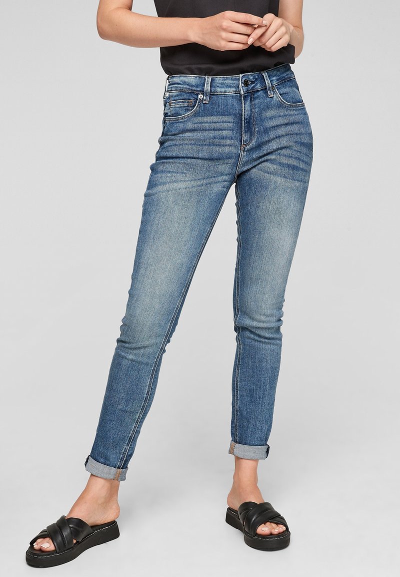 Jean en denim bleu ajusté avec une légère décoloration, cinq poches et des revers roulés, porté avec des sandales noires sans fermeture sur un fond neutre.