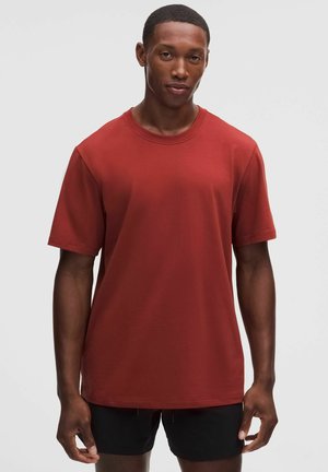 Junger Mann, der ein schlichtes rotes kurzärmeliges T-Shirt und schwarze Shorts trägt, steht vor einem schlichten hellen Hintergrund.