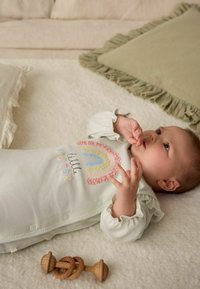 Ljusgrön babyoutfit med regnbågsdesign och texten "lilla syster"; textil med struktur, volangärmar, träleksak på mjuk filt.