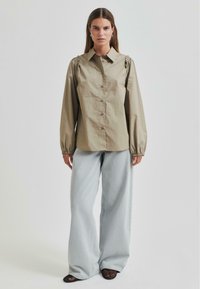 Smėlinis marškiniai su pūstais ilgais rankovėmis, derinami su šviesiai mėlynomis plačiomis kelnėmis. Medžiaga atrodo lygi, turi struktūruotą apykaklę ir rankogalius.