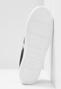 Svart sneakers med en texturerad tygöverdel, vit gummisula och blå accentrand på sidan. Har en platt, slät sula.