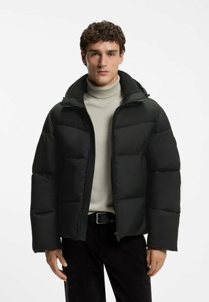 Junger Mann mit lockigem Haar trägt eine schwarze Steppjacke über einem hellbeigen Rollkragenpullover und steht vor einem einfarbigen weißen Hintergrund.