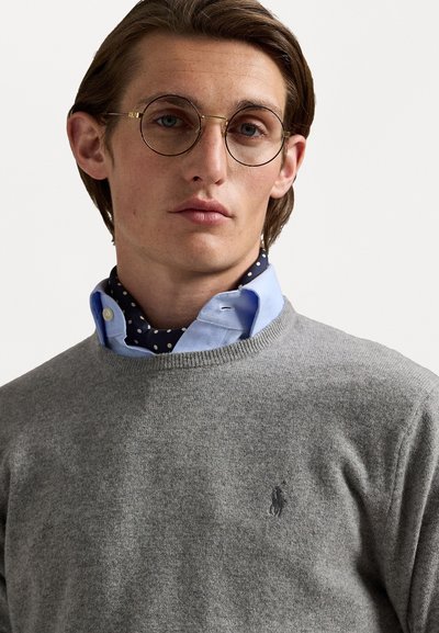 Un pull gris à col rond, superposé à une chemise bleu clair à boutons avec des pois blancs. Des lunettes rondes avec une monture dorée.