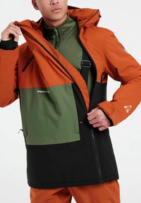 Orange und schwarze Jacke mit einem grünen Einsatz, ausgestattet mit einem Reißverschluss vorne, verstellbare Kapuze und Seitentaschen. Glatte Textur.
