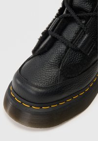 Bota de cuero negro con acabado texturizado, que cuenta con una suela de goma gruesa y costuras amarillas a lo largo del borde. Los cordones son visibles en la parte frontal.
