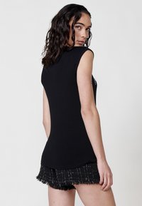 Musthave musta ilma varrukatega topp sileda tekstuuriga, kombineeritud musta frangi ja valgete ruutudega seeliku ning mudel pöördub veidi taha.