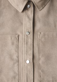 Beige corduroy overhemd met een gespitse kraag, twee borstzakken en zilveren drukknoopsluitingen aan de voorkant, met ribbelstructuur door het hele shirt.
