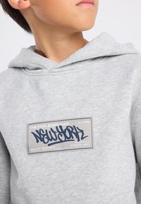 Sudadera gris con un parche frontal que dice "Nueva York" en azul marino, rodeado de un borde rectangular y texto. Tejido suave y diseño casual.