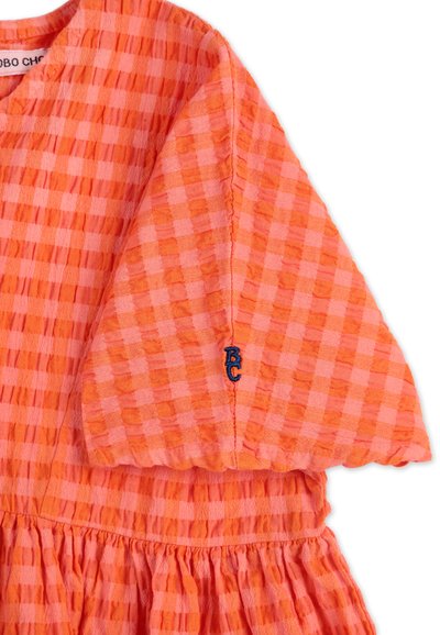 Orange och rosa rutigt klänningsärm med blå "BC" broderi och rynkad midjedetalj på vit bakgrund.