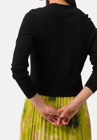 Černý pletený svetr s kulatým výstřihem a tříčtvrtečními rukávy, nošený přes plisovanou květinovou zeleno-žlutou sukni.