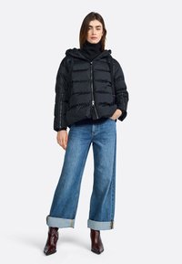Zwarte pufferjas met capuchon, ritssluiting en gequilt textuur, gecombineerd met lichtblauwe wijde jeans en puntige bruine enkellaarsjes.