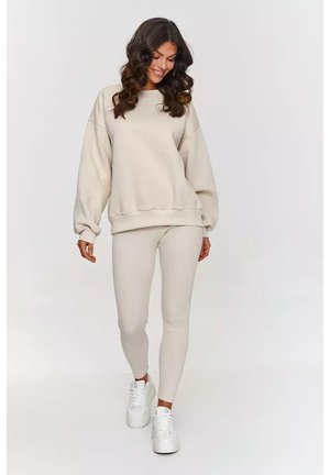 Beżowy, oversize'owy sweter z obniżonymi ramionami, zestawiony z prążkowanymi legginsami w pasującym kolorze. Białe sneakersy dopełniają look.