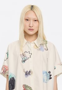 Camicia a maniche corte con bottoni in tessuto crema, caratterizzata da stampe astratte multicolori e forme delineate in nero sulla parte anteriore e sulle maniche.