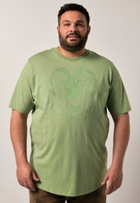 Camiseta de algodón verde con un estampado gráfico de un carnero rodeado de follaje decorativo. Mangas cortas y cuello redondo. Textura suave y corte relajado.