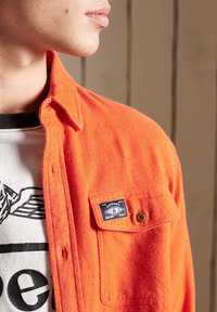 Kirkkaan oranssi flanellipaita, jossa on napillinen tasku ja sininen etiketti. Kangas on teksturoitu, näkyy graafisen t-paidan rinnalla paidan alla.